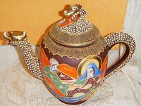 Front angled view of Satsuma porcelain teapot showing detailed enameled painted figures and dragon-shaped spout and handle with gilt accents