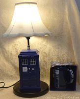 Photo of the TARDIS lamp illuminated, showing the blue base designed like a police call box and white fabric shade.