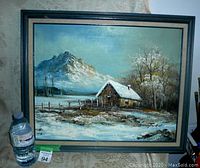 Framed oil painting on canvas showing a winter landscape with a snow-covered cabin, mountain, and trees, signed G. Whitman.