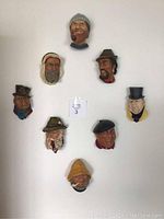 Photo showing all eight Boson head wall plaques arranged on a flat surface, each with distinct facial features, hats, and scarves.