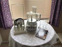 KitchenAid food processor set with base unit, attachments, and manual on a table with floral curtains in background.