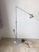 Silver adjustable IKEA floor lamp shown fully extended upright against white wall on carpeted floor, with power cord and transformer unit on floor.