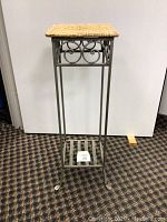 Front view of iron plant stand showing square woven top, decorative iron scrollwork, slender legs with curled feet and lower slatted shelf.
