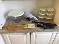 Wide view of all items including glass bowl with lid, knives, wooden spoons, small containers, pocket thermometer, and manual food chopper on countertop.