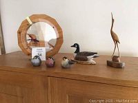 View of wooden crane bird figurine, Canada goose decoy, trio of small ceramic birds, and octagonal framed wooden mirror on wooden surface