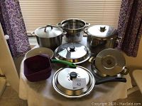 Photo showing mixture of pots, pans, lids, and a purple plastic strainer basket on a table
