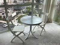 Side angle showing two white wooden folding chairs and round glass-top table near window with city view