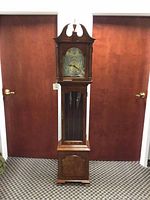 Front full view of the grandfather clock in wooden finish standing between two doors on carpeted floor.