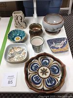 Photo showing eight varied pottery pieces arranged on a white table, including plates, bowls, vases, and a cup with decorative designs.