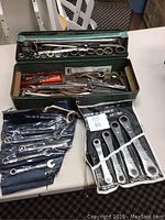 Photo showing an open green metal box with various socket wrenches, ratchets, and accessories along with two packs of wrenches, one fabric roll-up and one sealed pouch.