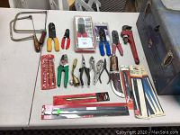 Photo showing assorted hand tools including crimpers, hacksaws, blades, a pipe wrench, and more, laid out on white table