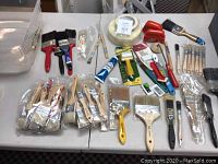 Top-down view of multiple paint brushes, rollers, masking tapes and paint accessories arranged on a table.