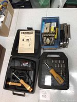 Overview photo showing Black & Decker heat gun, driver set case, staple guns, and hardware box on table.