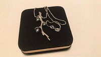Photo of sterling silver necklace with a branch-shaped pendant attached to a delicate chain, showing three dark blue pearls. Necklace is placed on a black fabric jewelry display.