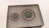Two sterling silver floral brooches displayed in a gray box: one larger with green and red stones and one smaller with blue and red stones.