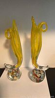 Front-side view of two yellow blown glass vases with clear round bases containing colorful glass ribbons, showing side handles and twisted forms.