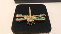 Dragonfly brooch with gold-tone metal, green enamel wings, and clear rhinestones on black velvet background.