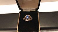 Sterling silver ring with blue lapis stone shown inside an open black and gold-trimmed ring box with beige interior.