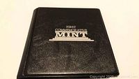 Black leatherette case with gold embossed 'First Commemorative Mint' text on the cover.