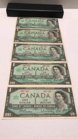 Five Canadian one dollar bills from 1967 laid flat, with a black case above them.
