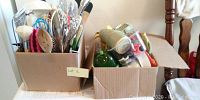 Two cardboard boxes containing assorted cooking utensils such as spoons, whisks, ladles, spatulas, peelers, jar opener, in mixed materials and colors.