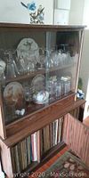 Photo showing upper glass door cabinet and lower wooden storage with doors. Glassware and decorative plates visible inside upper cabinet but contents excluded.