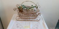 Large decorative metal sleigh with handle and faux greenery inside, intricate scroll design, metallic finish.