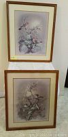 Two framed prints hang on or rest against a white wall. Depict birds on floral branches with soft pastel backgrounds. Both are signed by the artist.