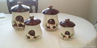 Photo showing four ceramic canisters with mushroom designs and brown lids on a table.