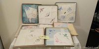 Photo of seven boxed and loose vintage handkerchiefs arranged on a table, showing various embroidered floral patterns, scalloped edges and pastel colors.