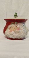 Photo showing a ceramic Santa Claus cookie jar with a red hat lid topped with a small green Christmas tree handle.