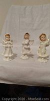 Three small angel figurines in white robes with short hair, standing on a flat surface against a plain backdrop.