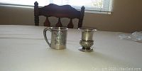 Photo of a vintage silver mug with ornate handle and engraved designs, next to a vintage silver vase with decorative ribbed base, both placed on a white table.
