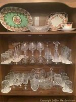 Wide view of shelves displaying assorted glassware including wine glasses, champagne flutes, cocktail glasses, cut-glass bowls and a teacup with saucer, plus two decorative trays on top shelf