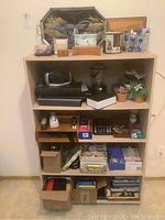 Full view of wooden bookcase with shelves holding multiple items including electronics, plants, media, and boxes.