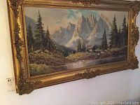Full view of the signed landscape painting with ornate gold frame showing mountain peaks, pine trees, cabin and reflective water.