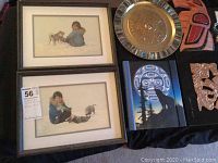 Two framed Inuit prints, engraved metal plate, small canvas print and painted wooden carving displayed on black fabric