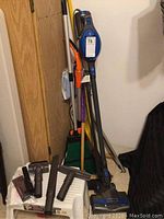 Rocket cordless vacuum cleaner leaning against wall with broom and dustpan behind, and step stool at base. Various vacuum attachments placed on step stool.