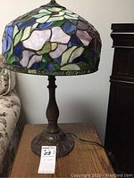 Full view of Tiffany style table lamp on wooden surface near beige floral couch, showing stained glass shade and metal base.