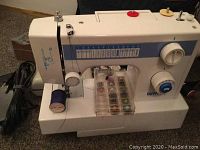 Front left angle photo of the white zig zag sewing machine with spool of blue thread and bobbins case visible.