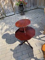 View of the two-tier wooden table showing the top and bottom tiers with scalloped edges and the tripod legs outdoors on stone pavement.