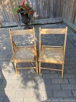 Pair of wooden folding cane woven chairs side by side outdoors on stone pavement.
