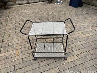 Metal bar cart with black metal frame and white perforated metal shelves photographed from an angle showing the structure and wheels.