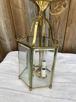 Front and side view of vintage solid brass lantern light fixture showing six clear glass panels, three white candle-style bulb holders inside, brass feet at base, and curved brass supports at top with hanging canopy.
