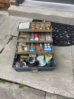 Open blue fishing tackle box on stone steps showing layered compartments with assorted fishing lures, hooks, spools of line, and vintage fishing accessories.