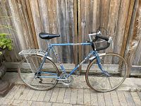 Full side profile of a vintage blue Raleigh road bike against a wooden fence showing frame, wheels, handlebars, saddle, and rear rack.