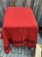 Red wool blanket draped over a table outside, showing overall color and black border along edge.
