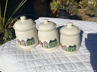 Three ceramic canisters arranged on a quilted white surface outside, showing their decorative house design.