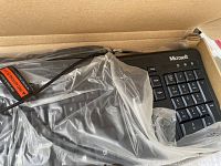 New Microsoft wired keyboard partially visible, wrapped in clear plastic inside cardboard box.