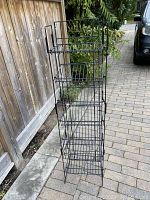 Front view of black metal wire stand with seven shelves displayed outdoors on a patio next to a wood fence.
